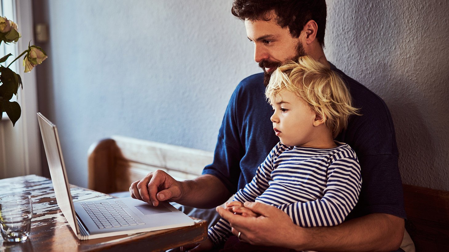 A father sits with his infant son in his lap as they look at a laptop screen