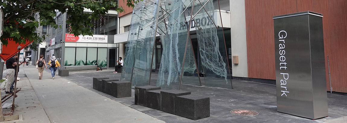 Sidewalk view of Grasett Park with a storefront and a vertical mesh art installation.