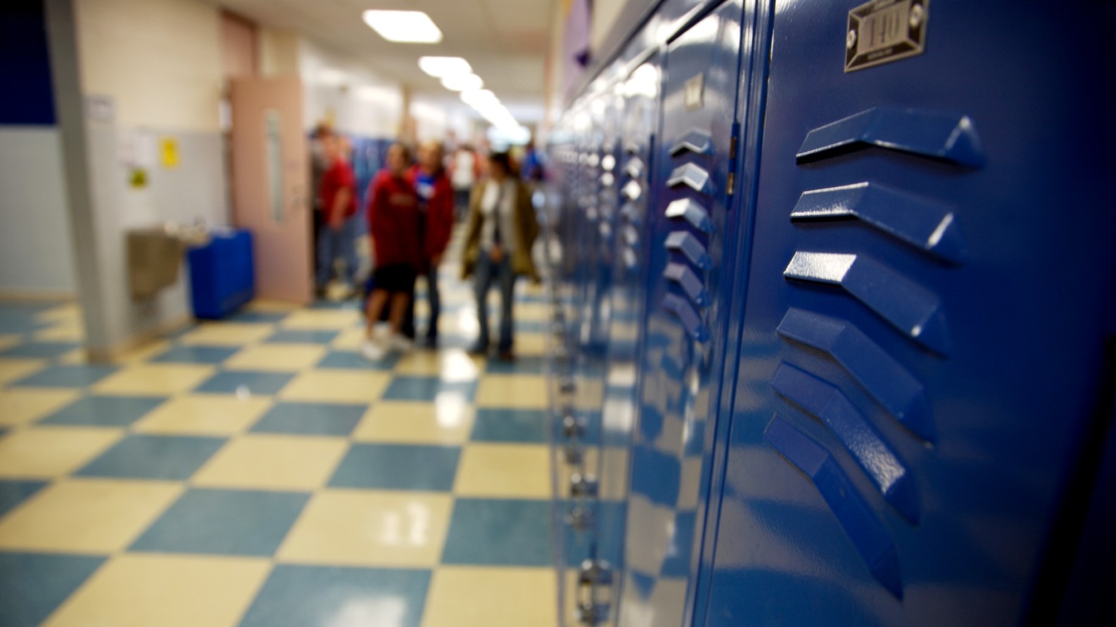Blue lockers line a school hallway with a blue-and-white checkered floor, while several students walk in the background out of focus
