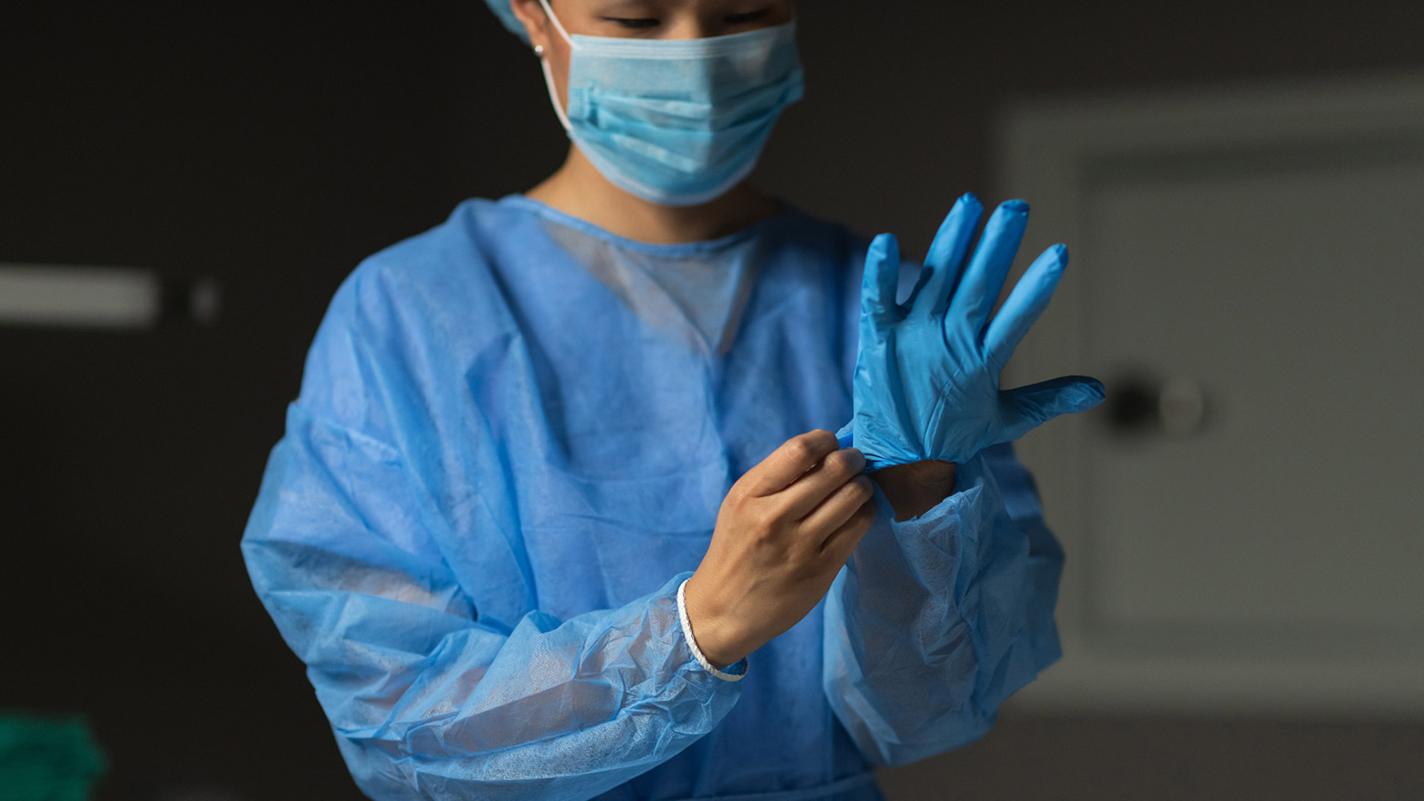 A female doctor wears a medical mask and a surgical gown while putting on medical gloves