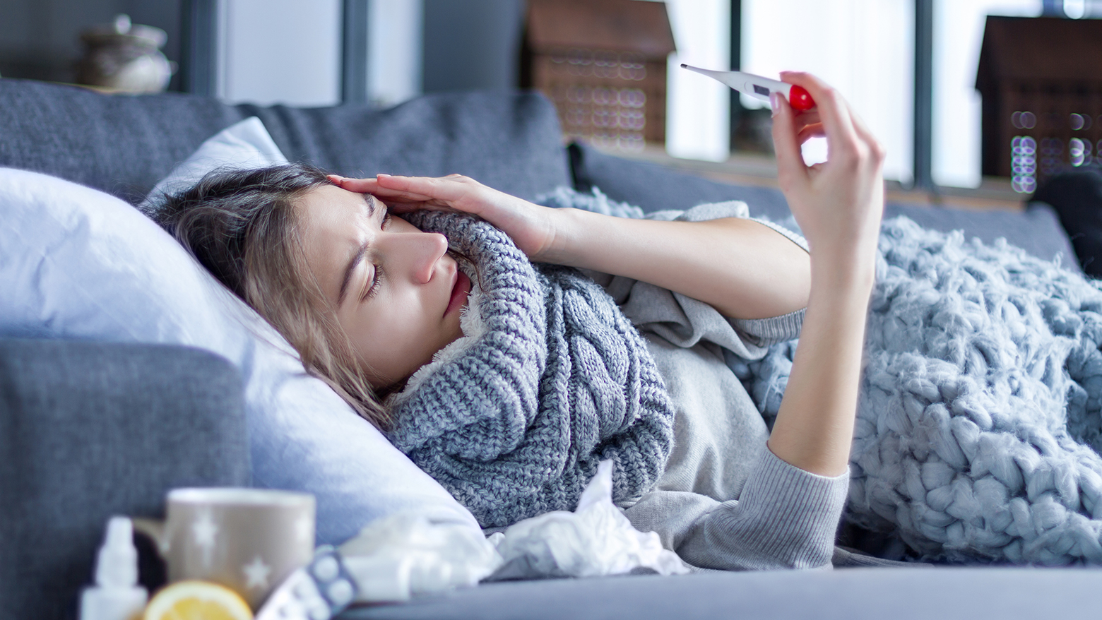 A woman checks her temperature with a thermometer as she lays on a sofa