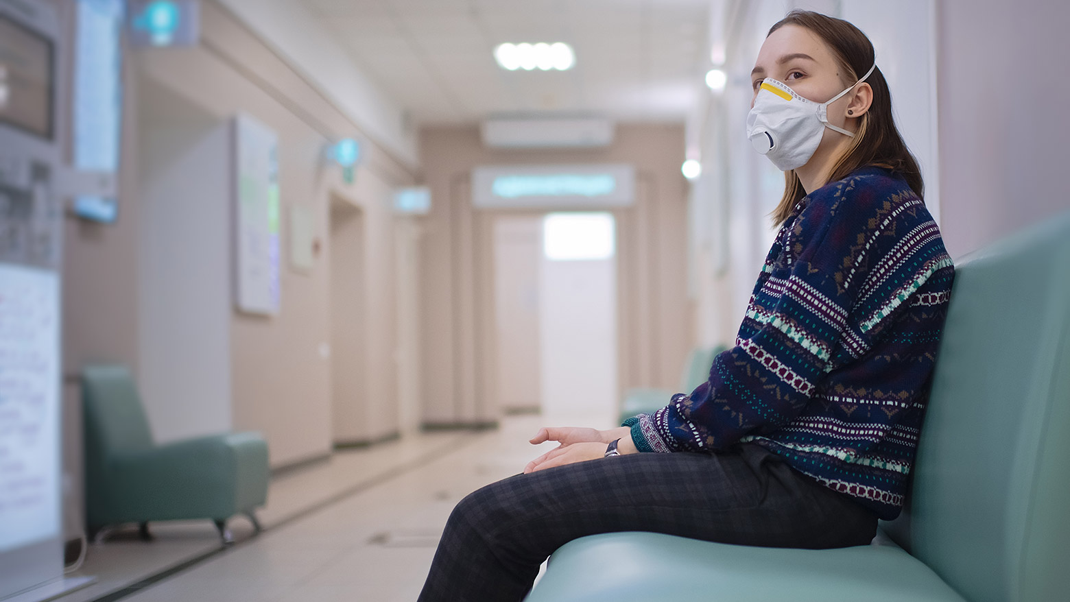 A female patient sits in a waiting room while wearing a medical mask