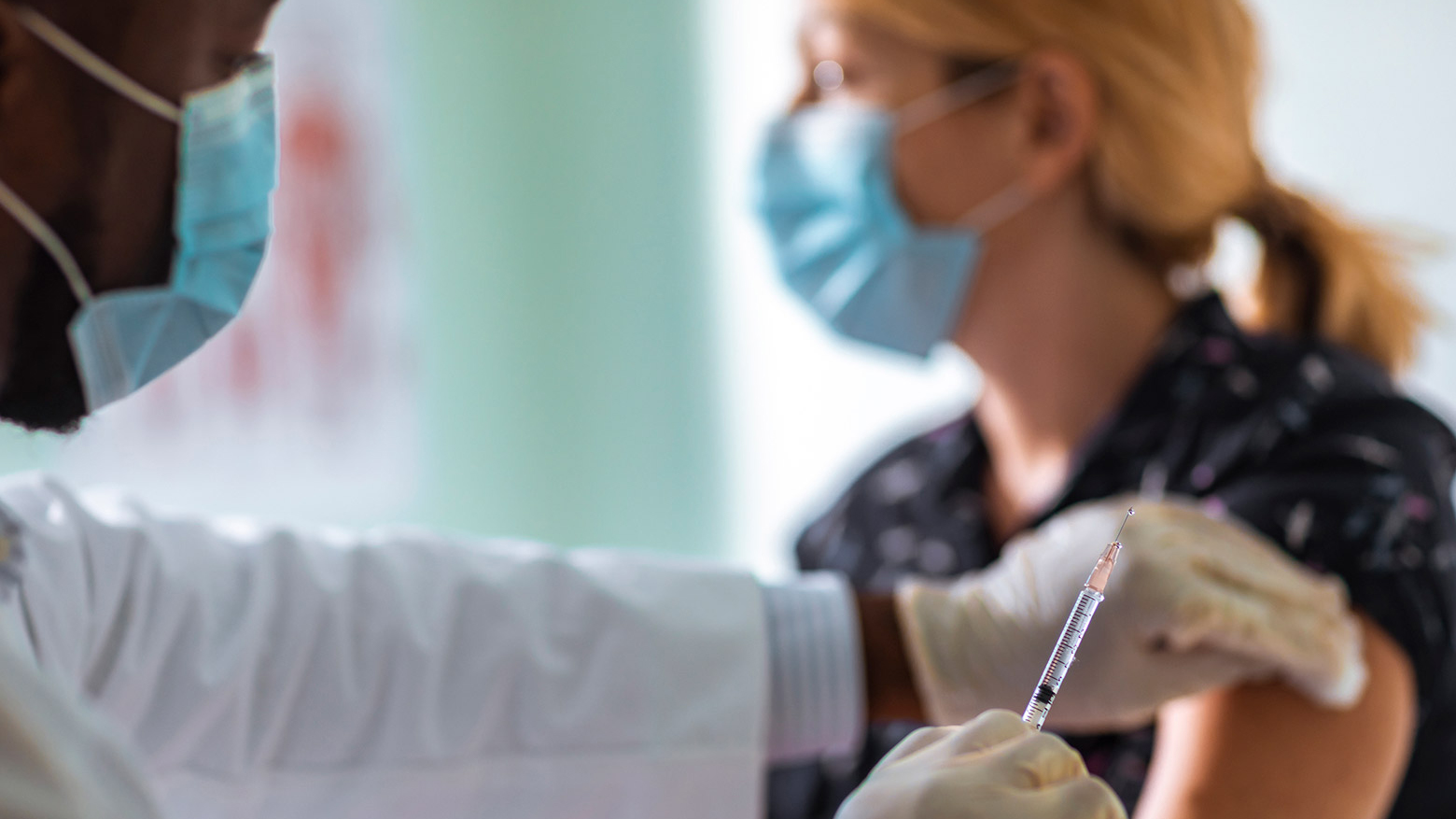 A male doctor wears a medical mask and gloves while administering a needle injection to a female patient in a medical mask