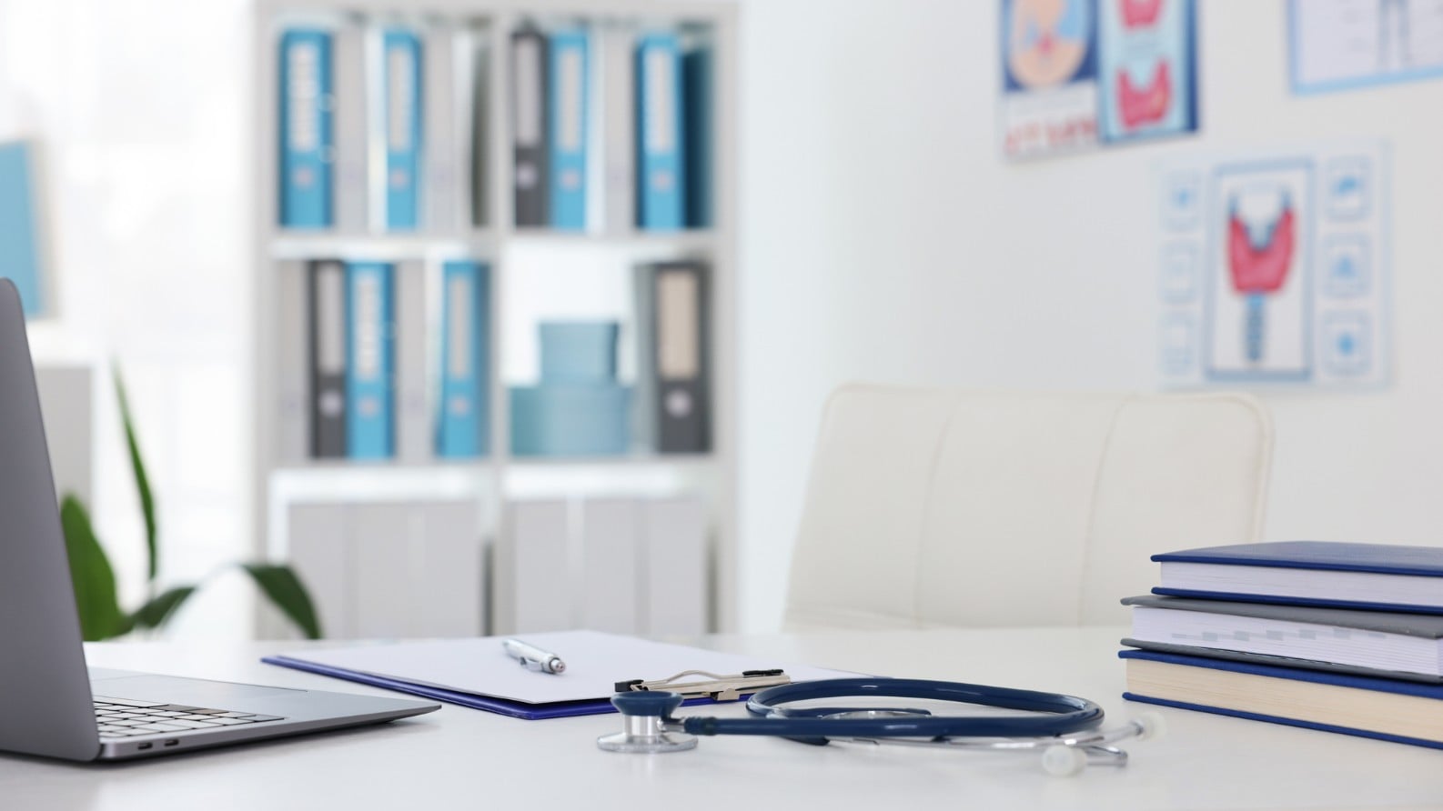 Desk setup in a medical office with a laptop, clipboard and pen, stethoscope, and stacked books, with shelves of binders and medical diagrams visible in the background.