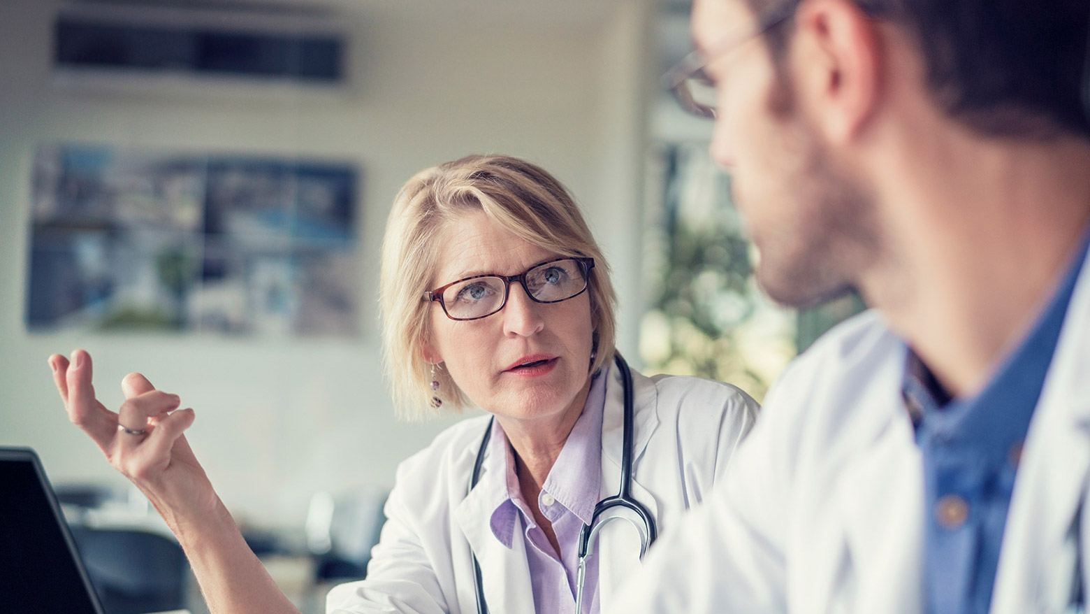 A male and female doctor sit beside each other at a desk while conversing