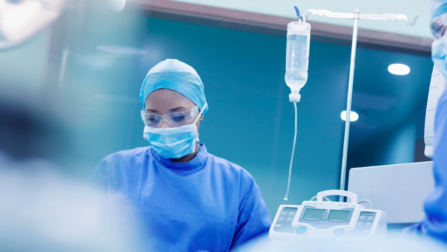 A female doctor wears medical scrubs and a mask while standing next to an IV fluid bag