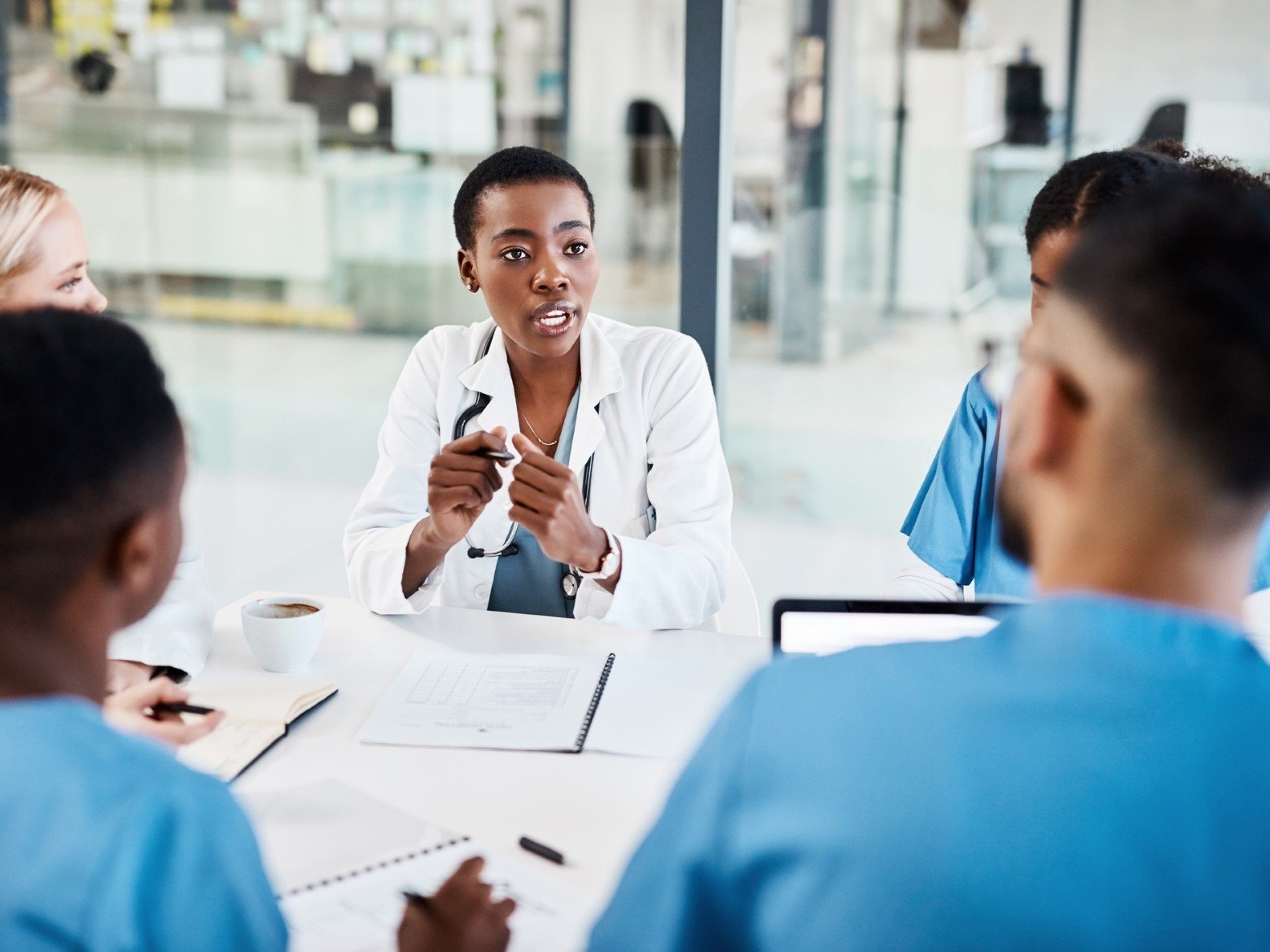 A group of healthcare professionals in blue scrubs and a white coat engaged in a discussion around a conference table.