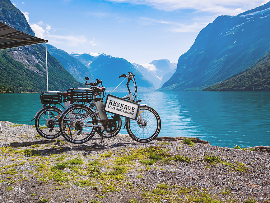 Two bicycles parked on a gravel path beside a large, serene lake surrounded by mountains. One of the bicycles has a sign attached to it that reads 'RESERVE YOUR RETIREMENT.' The scene is set in a picturesque location with clear blue water and lush green mountains under a partly cloudy sky.