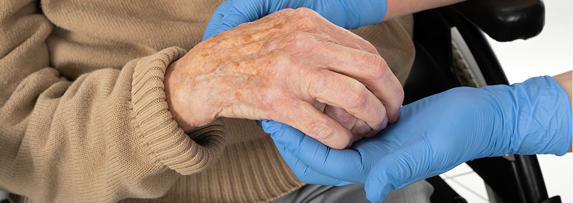 A healthcare worker wearing gloves holds the hands of an elderly patient