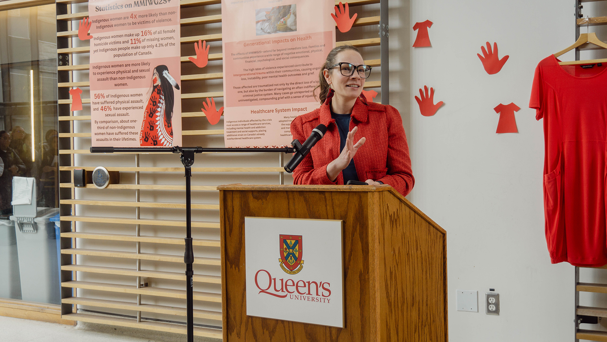 Dr. Jamaica Cass speaks from a Queen’s University podium in front of MMWG2S+ (Missing and Murdered Indigenous Women, Girls, and Two-Spirit people) awareness posters and red dress cutouts.