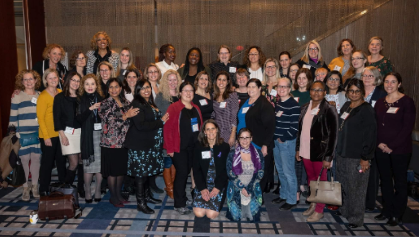 A large group of people posing for a group photo in an indoor setting, possibly at an event or conference. Some individuals are standing while others are kneeling, and many are wearing name tags. The background includes a curtain and part of a staircase.