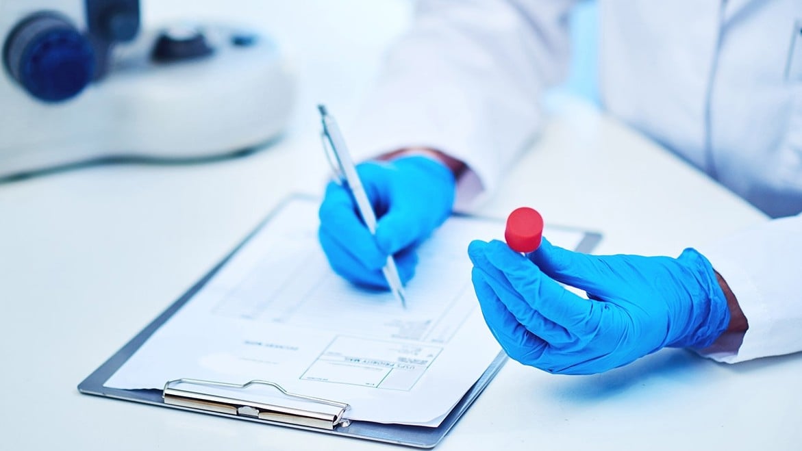 A pair of hands with blue protective gloves handles a vial in a laboratory.