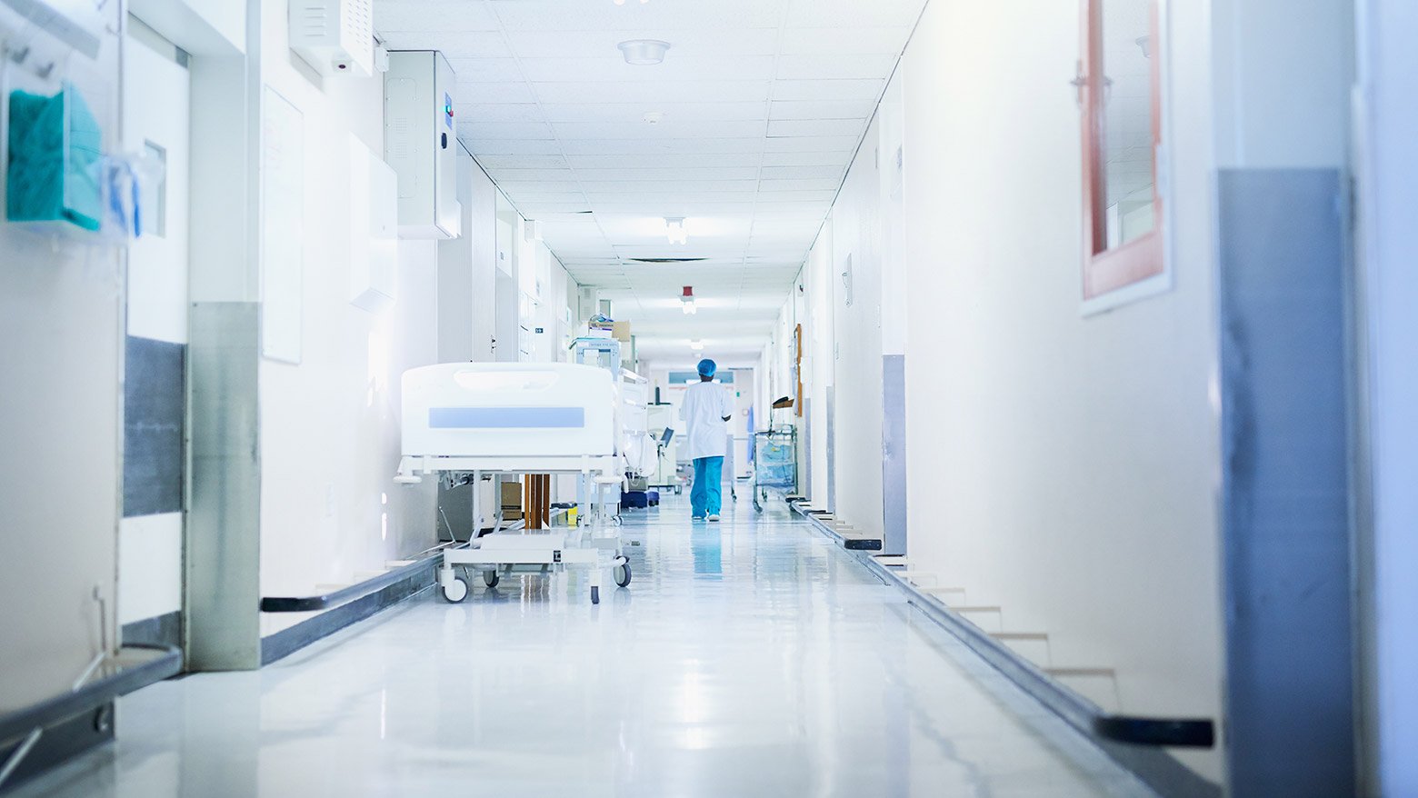 A long, clean hospital corridor with bright lighting. Medical equipment and supplies are along the walls, and a person in medical attire is walking down the hallway. The floor is shiny and reflective, indicating cleanliness.