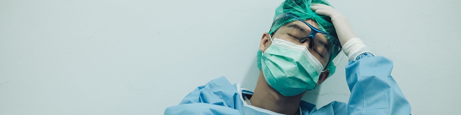 Tired doctor in scrubs and mask leaning against wall