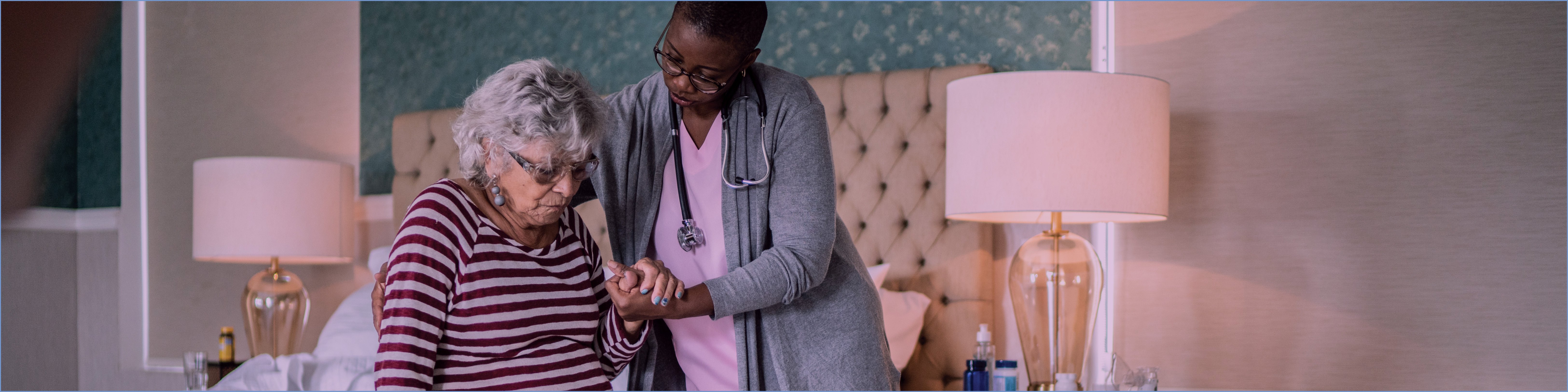 A professional wearing a stethoscope holds the hand of an elderly woman in the act of helping her get up from a bed.