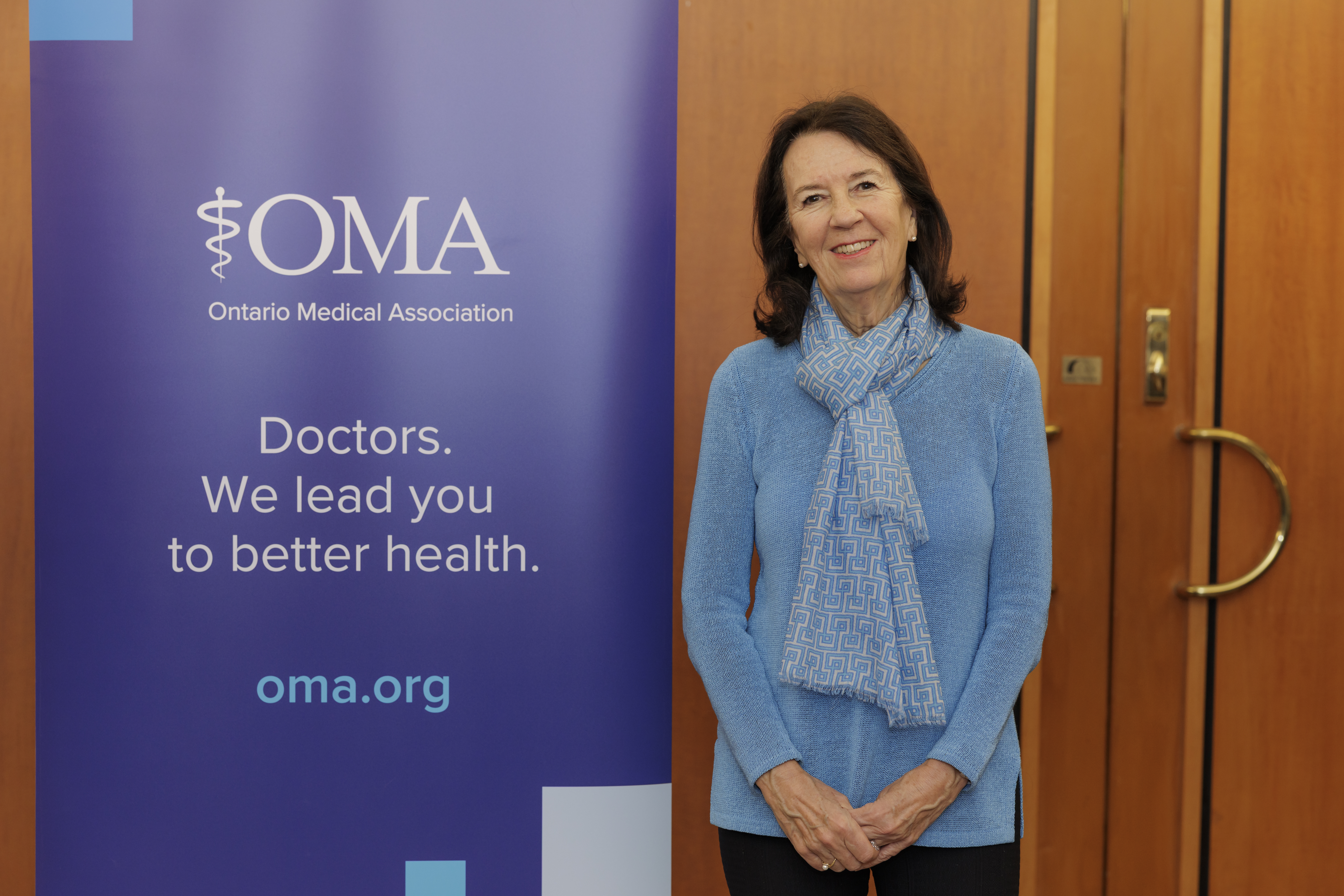 Dr. Jane Charters stands in front of wooden doors and alongside an Ontario Medical Association-branded banner that reads, 'Doctors. We lead you to better health.' 