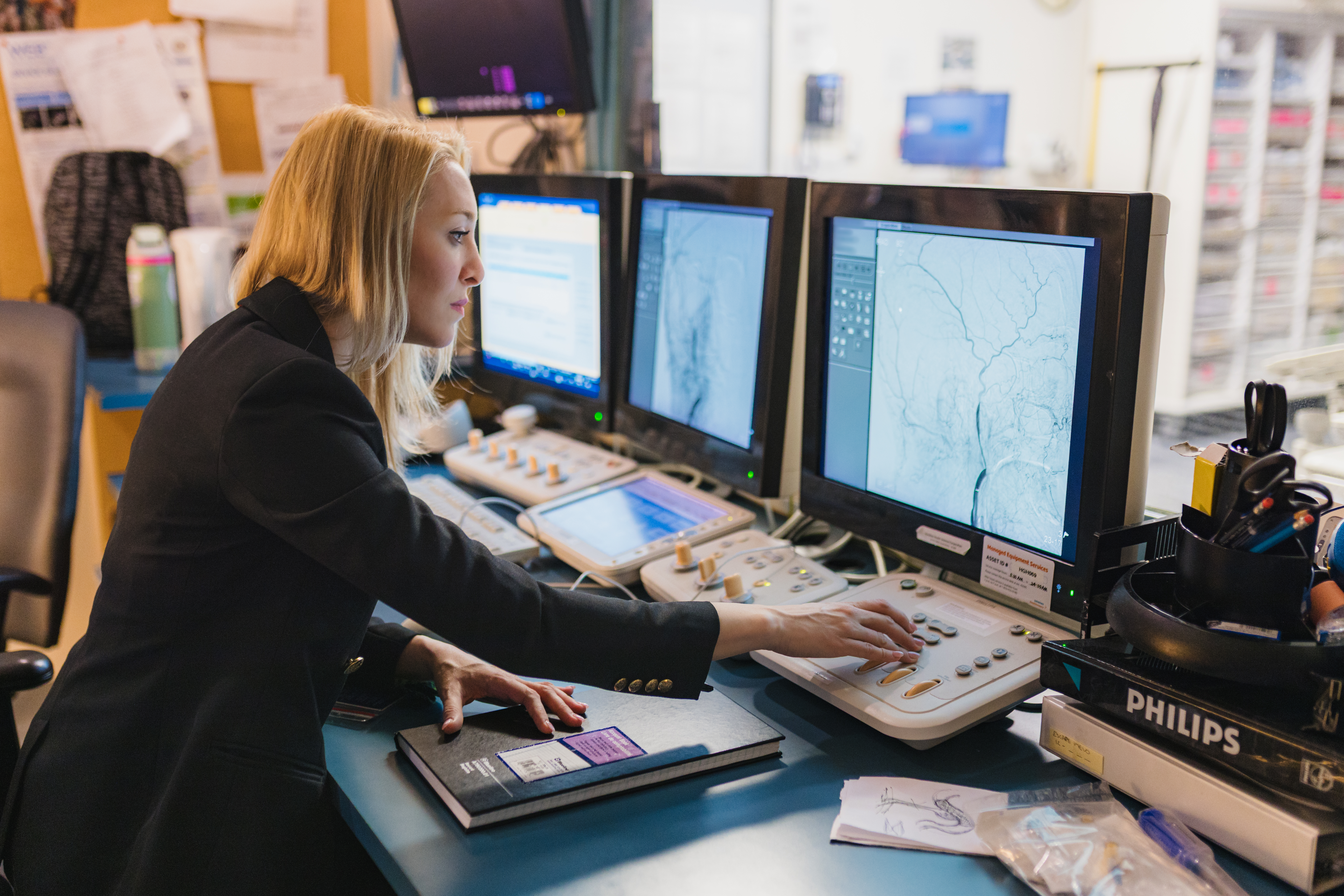 Dr. Luciana Catanese looking closely at brain angiograms on multiple computer monitors in a clinic settings.