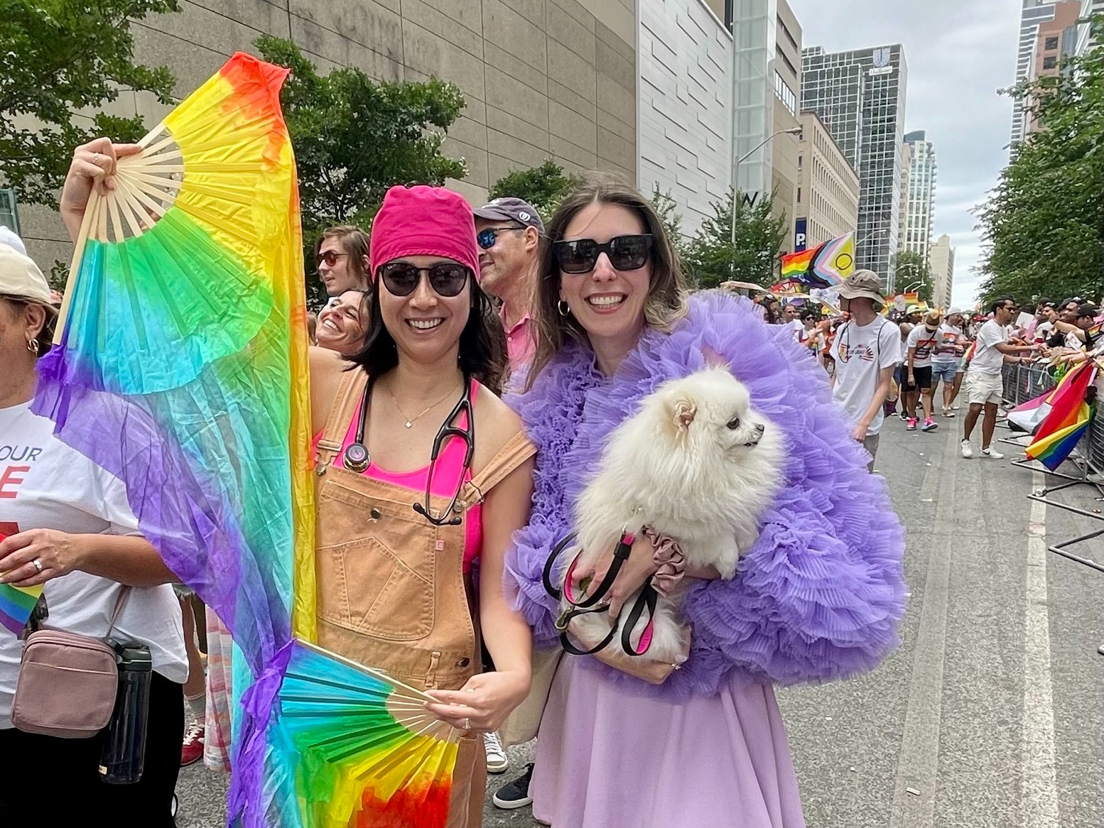 Two women stand together on a downtown Toronto street smiling. The woman on the left wears beige overalls, a pink t-shirt, a pink bandana, black sunglasses and holds rainbow fans, while the woman on the right wears a lavender skirt, dark purple frilly sweater and dark sunglasses, holding a small white fluffy dog.