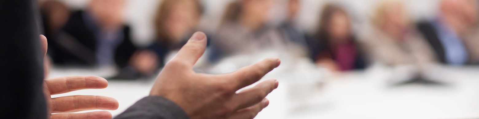 A person talks with their hands as a crowd, in the background, listens.