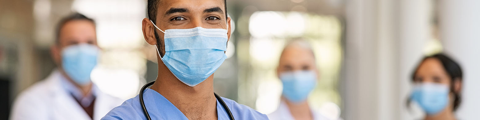 male doctor wearing scrubs and a mask in the foreground with three other doctors in masks behind him