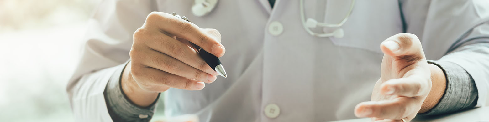 A health care worker in a lab coat with a stethoscope and a pen in hand.