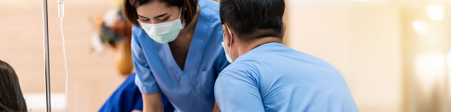 A health-care worker in a mask and scrubs leaning over to help a patient.