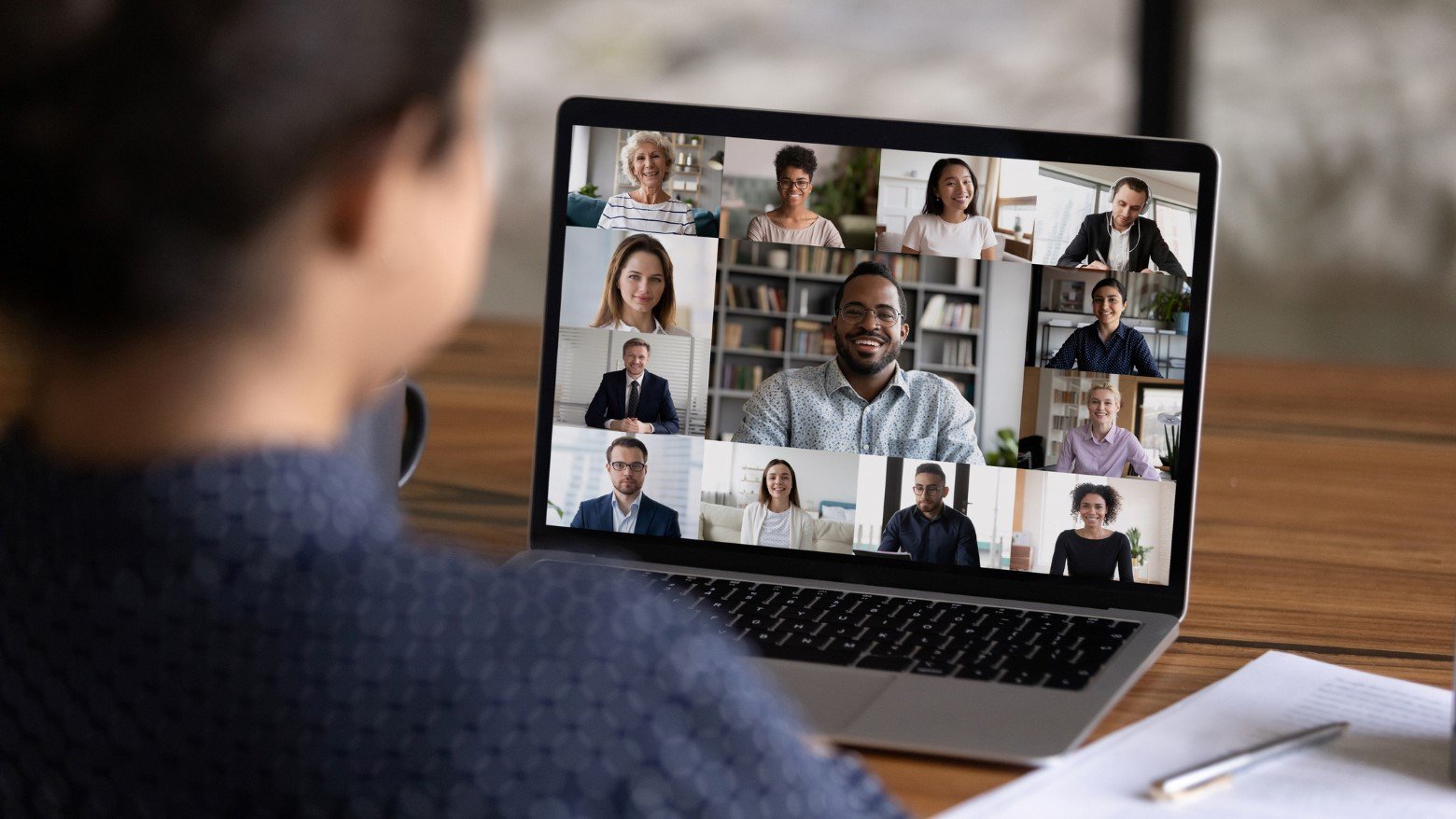 A person sits at a desk, engaged in a video conference on their laptop displaying multiple participants in a grid layout.