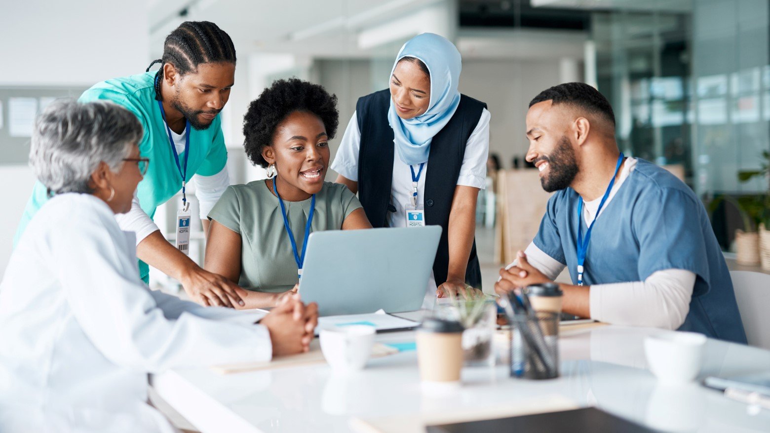 A diverse group of healthcare professionals collaborates around a laptop in a modern office setting, engaged in discussion.