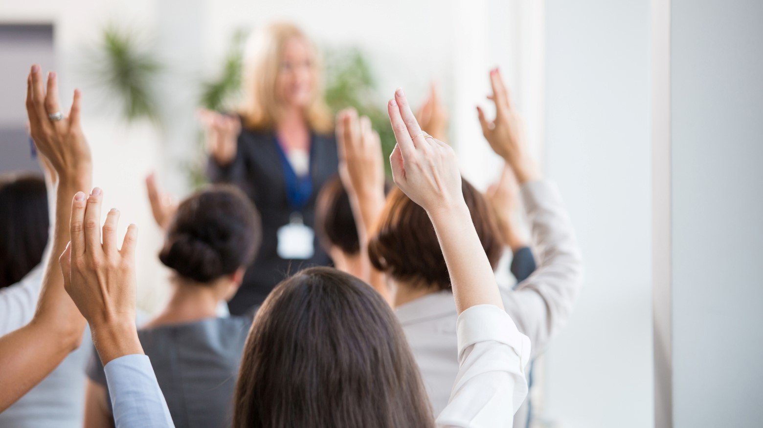 A group of people with raised hands, actively engaged in a seminar or discussion, facing a speaker in a bright, modern environment.