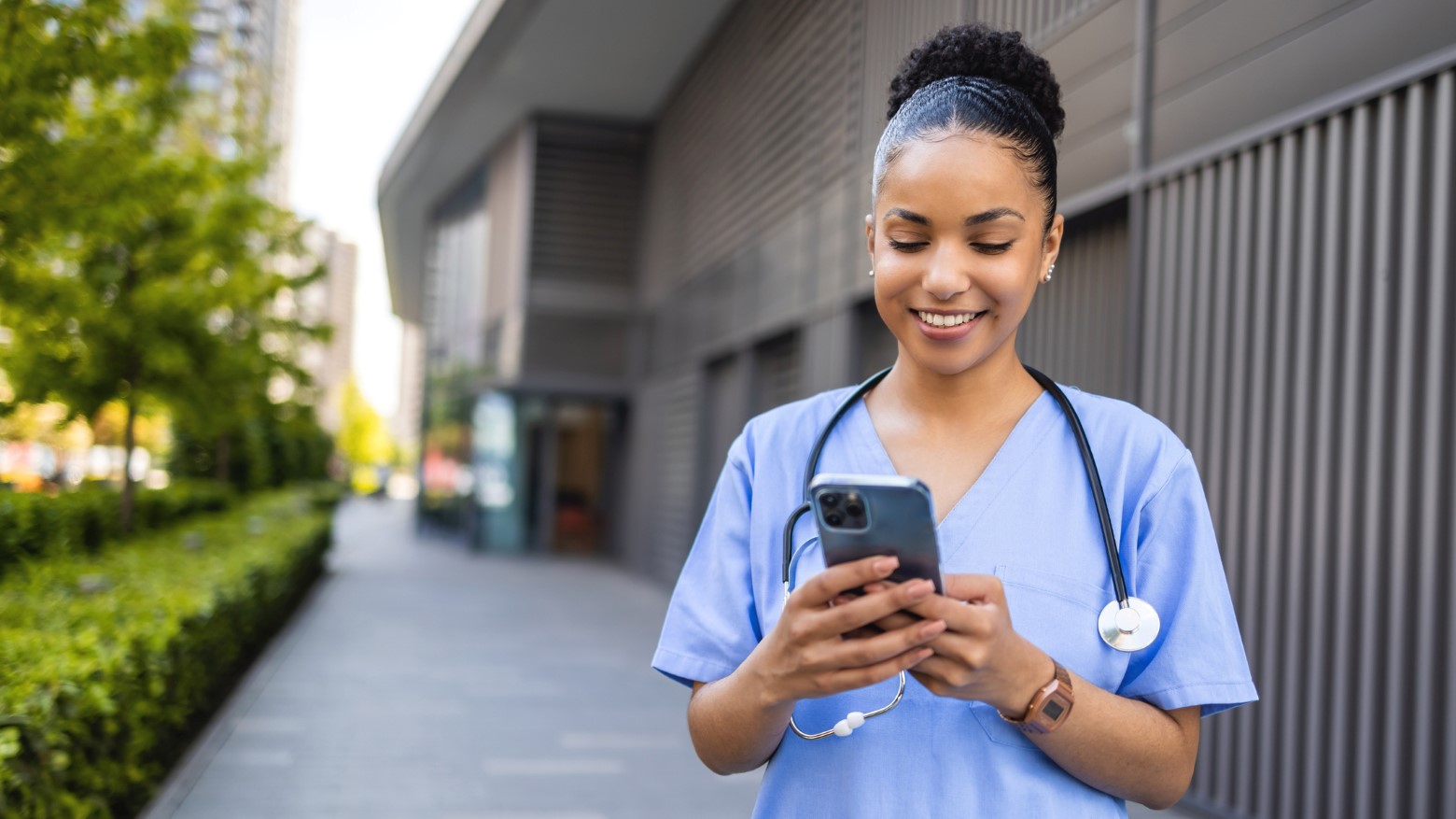 A healthcare professional in blue scrubs holds a smartphone, standing outdoors beside greenery and modern buildings.