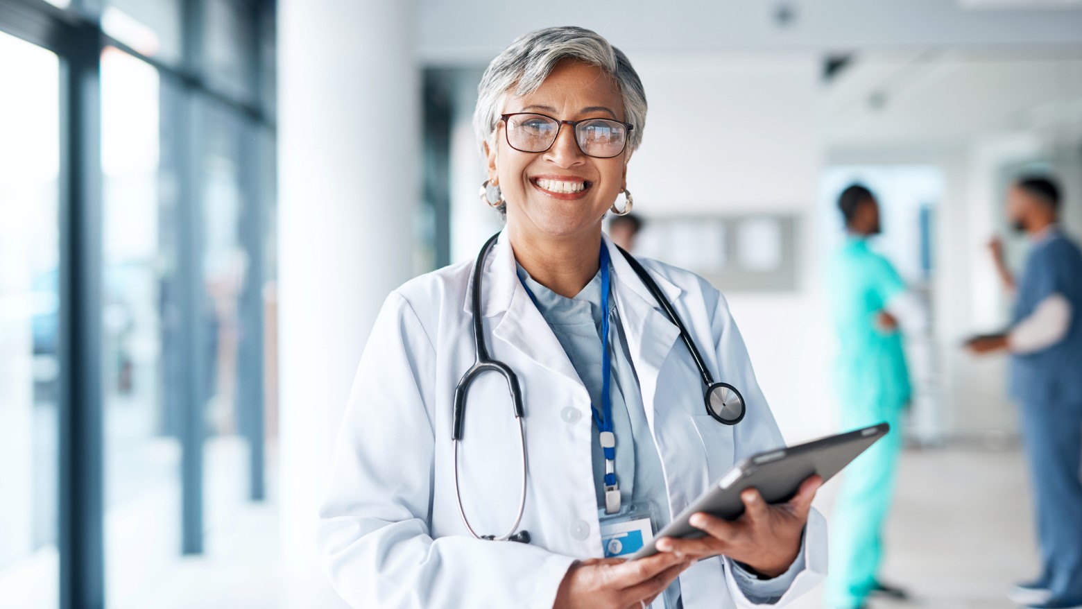 A healthcare professional in a white lab coat and stethoscope holds a tablet, with hospital staff visible in the background.