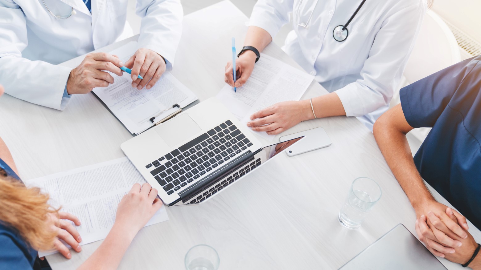 A group of healthcare professionals in white coats discussing paperwork and using a laptop during a meeting at a conference table.