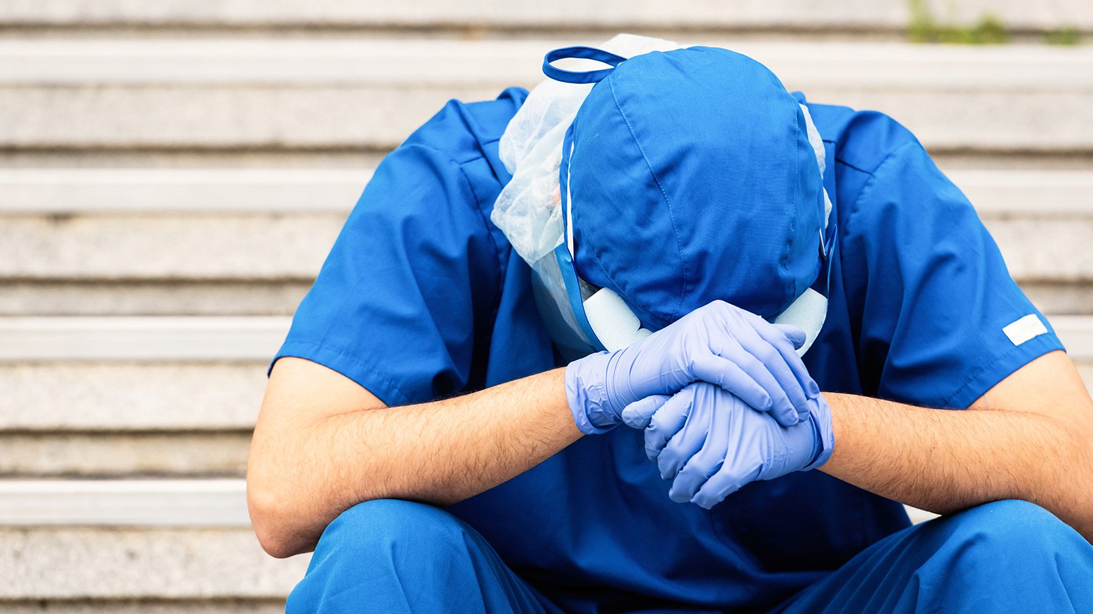 Male health care worker in scrubs and PPE with head down in hands.