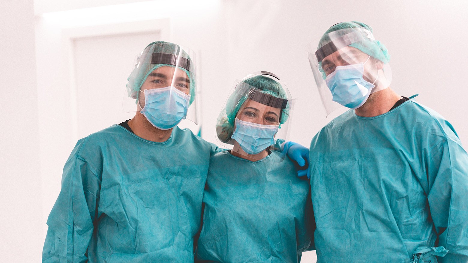 Three health-care workers standing side-by-side in full personal protective equipment (masks, face shields and gowns)
